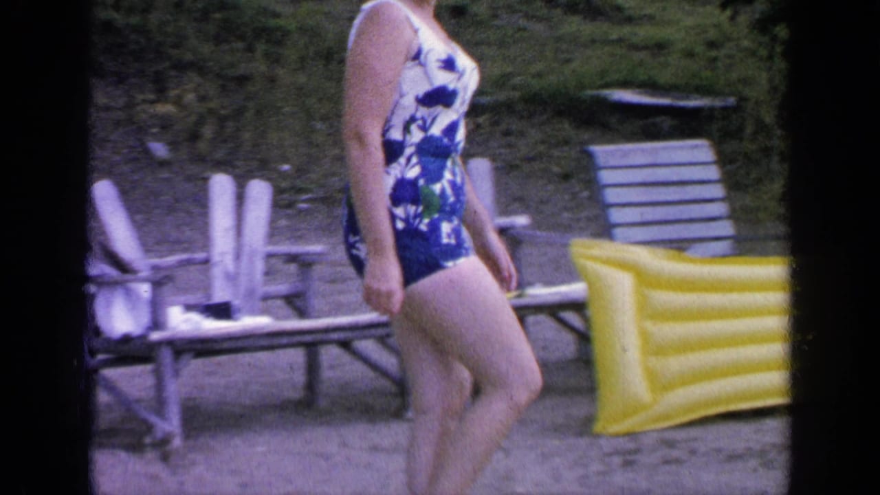 Woman in a floral swimsuit walks beside lake chairs and a yellow inflatable float.