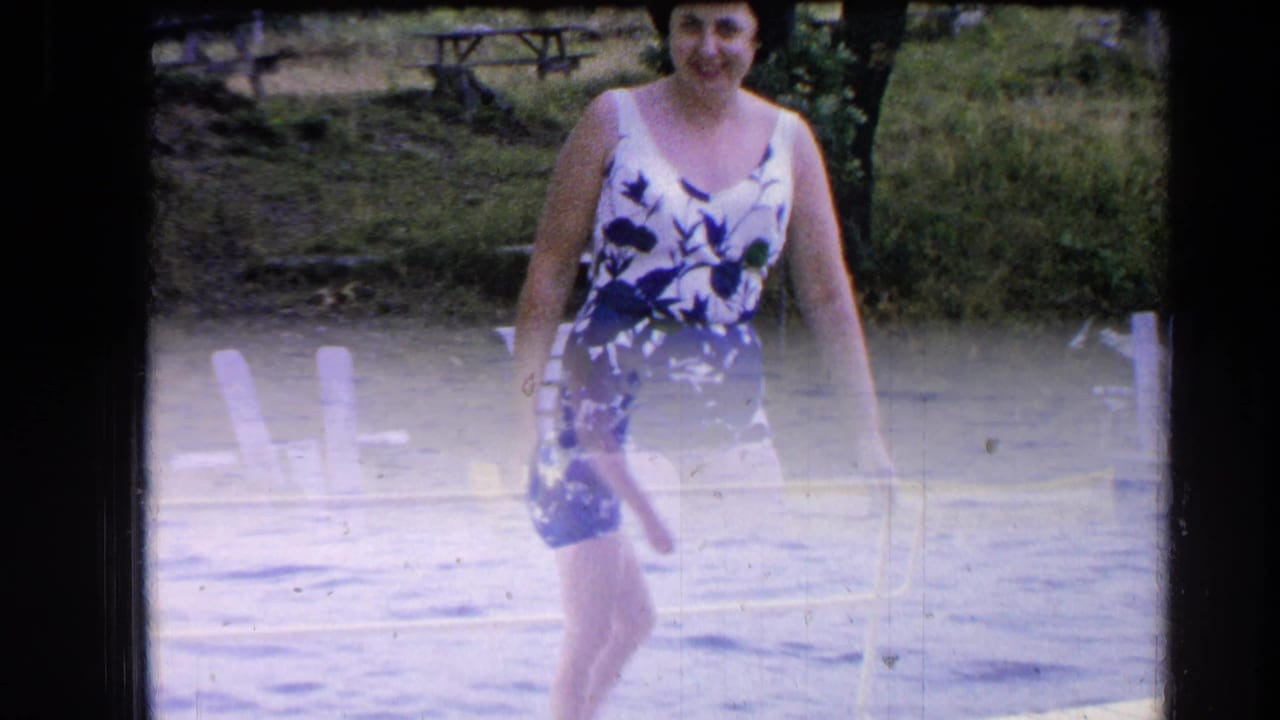 Woman in a floral swimsuit faces the camera while walking near the lake edge.