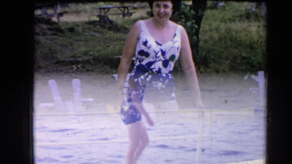 At Greenwood Lake in 1964, the woman in a floral one-piece swimsuit walks toward the camera near water and shoreline chairs.