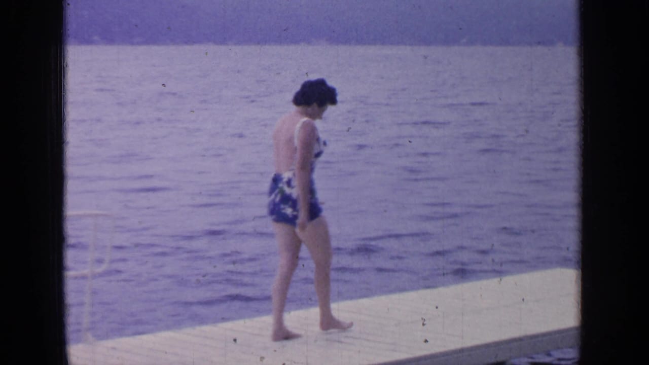 At Greenwood Lake in 1964, an adult woman in a blue and white one-piece swimsuit walks barefoot on a wooden platform by the water.