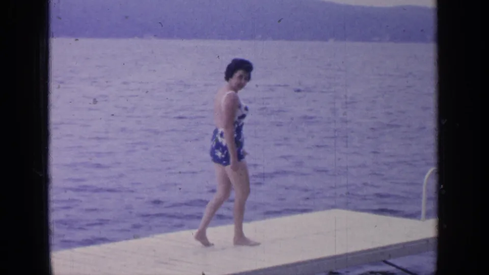 The woman walks near the edge of the Greenwood Lake platform, with open water and low hills visible across the background.