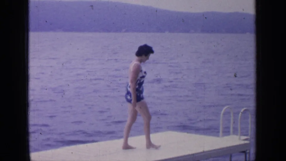 The woman approaches the platform end near a metal ladder, looking down toward the water at Greenwood Lake in this 1964 home-movie frame.
