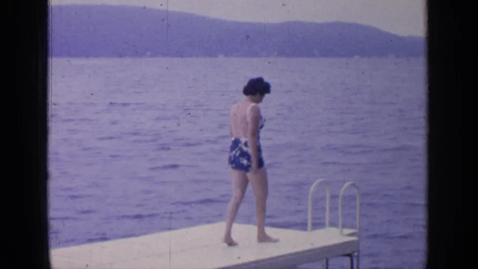 Near the end of the Greenwood Lake platform, the woman pauses beside the metal ladder with the water stretching behind her.