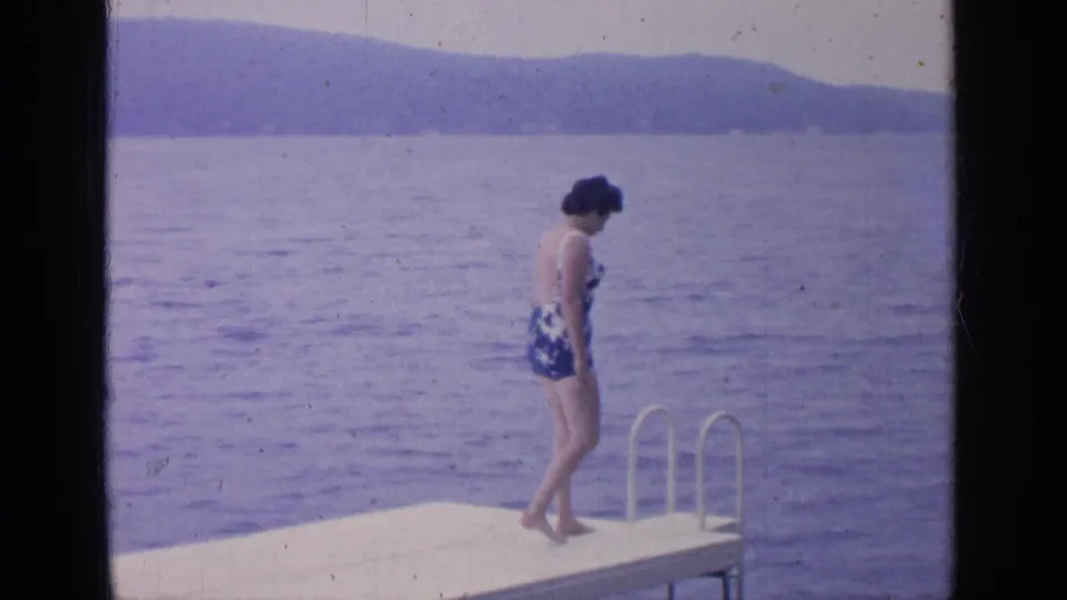 At Greenwood Lake in 1964, a woman in a blue and white one-piece swimsuit stands on a dock near a metal ladder.