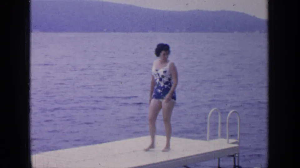 At Greenwood Lake in 1964, the woman stands on the dock near the metal ladder, looking to one side across the lake.