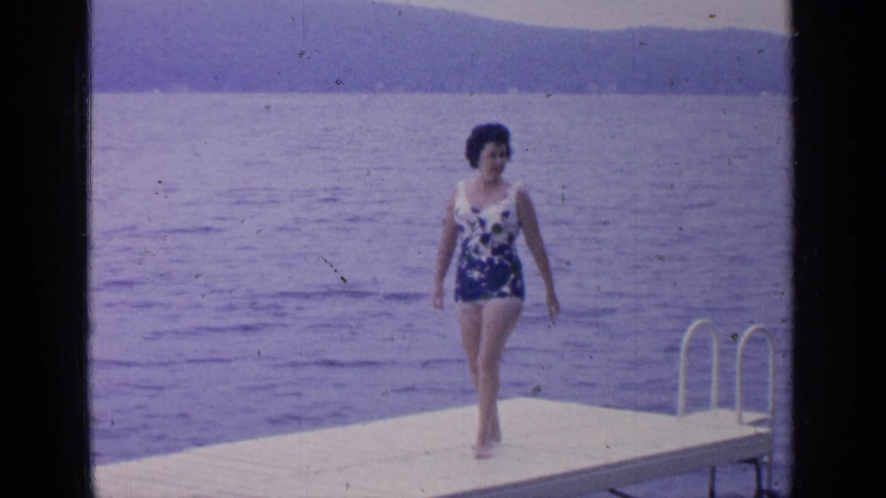 At Greenwood Lake in 1964, a woman in a blue-and-white swimsuit walks barefoot on a white dock with water and hills behind her.