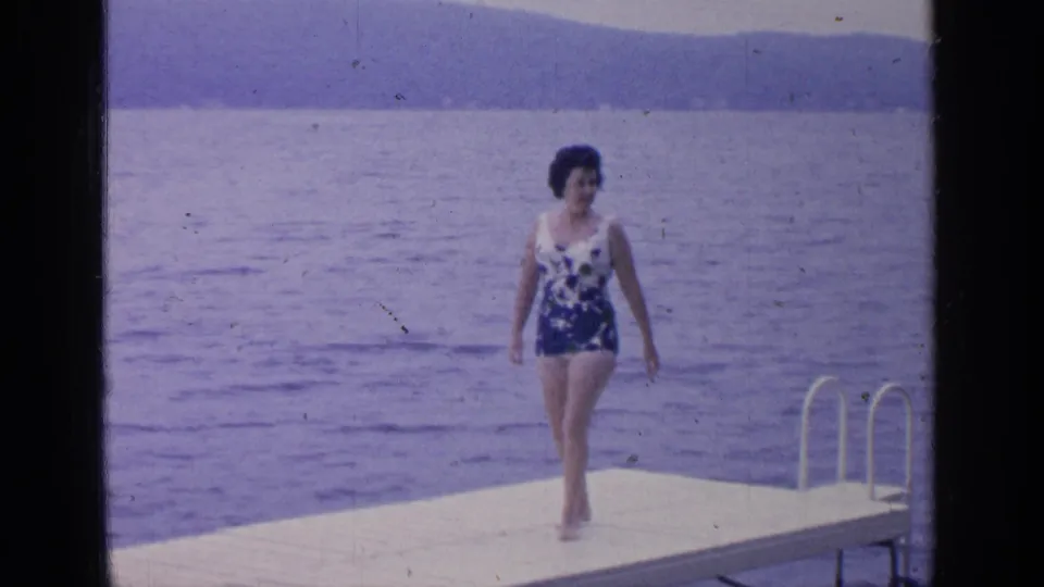 At Greenwood Lake in 1964, a woman in a blue-and-white swimsuit walks barefoot on a white dock with water and hills behind her.