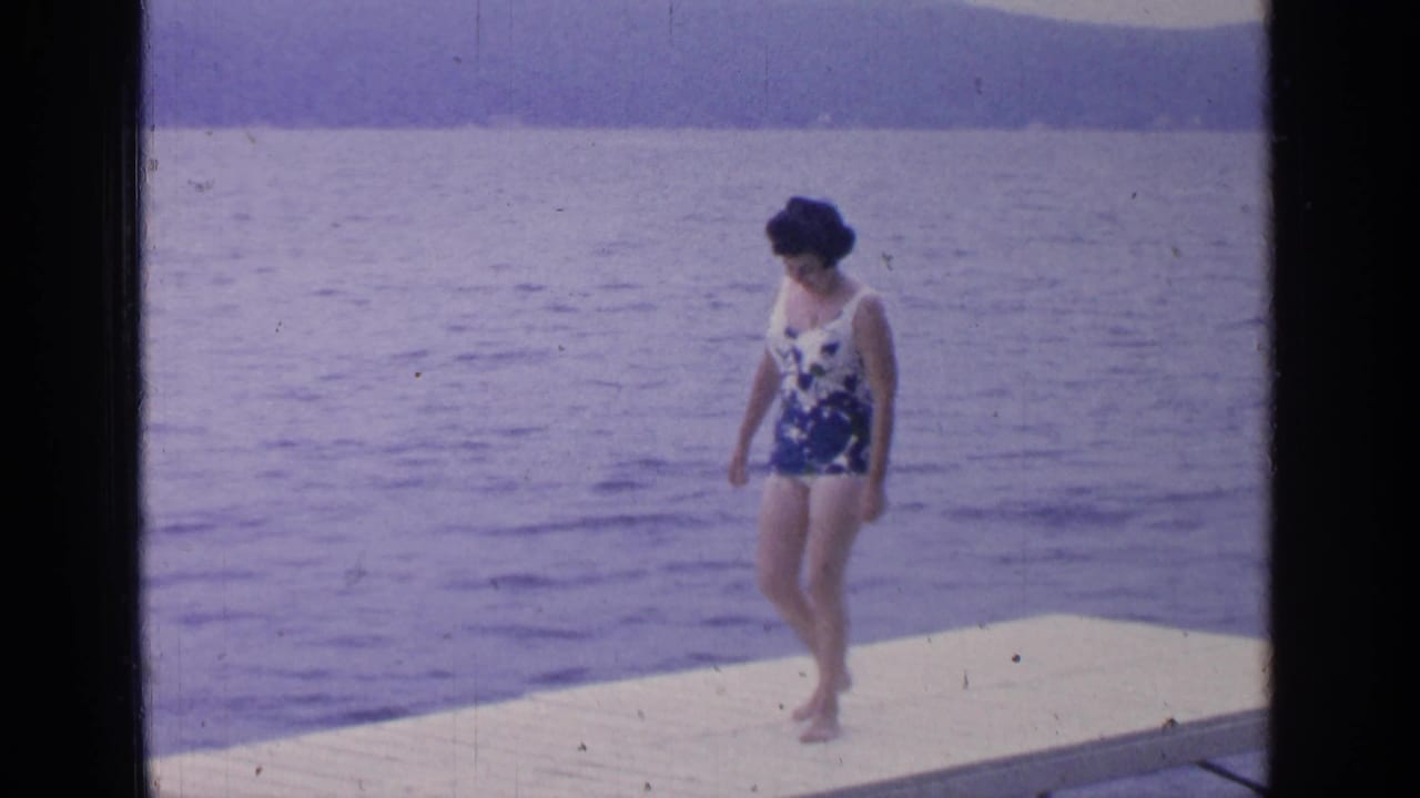 The woman continues across the white dock at Greenwood Lake, with calm water and distant hills visible in the 8mm frame.