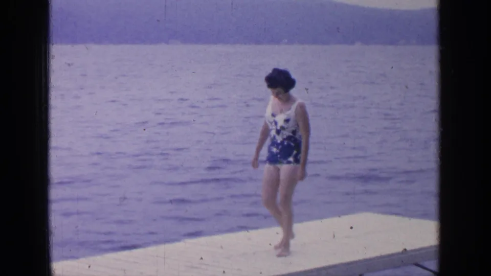 The woman continues across the white dock at Greenwood Lake, with calm water and distant hills visible in the 8mm frame.