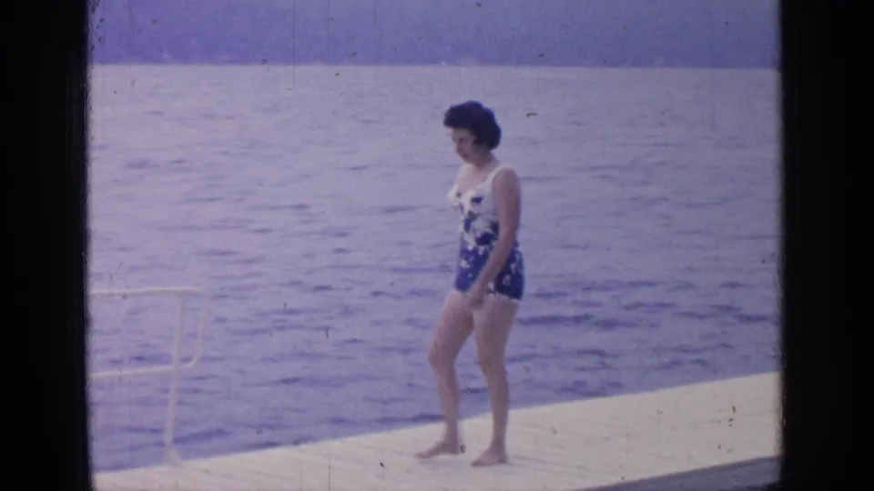 A woman in a floral one-piece swimsuit appears near the dock edge at Greenwood Lake, with water filling most of the background.