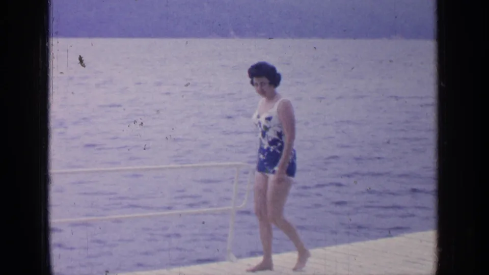 The woman walks beside a low white rail on the Greenwood Lake dock, framed by blue lake water and distant hills.