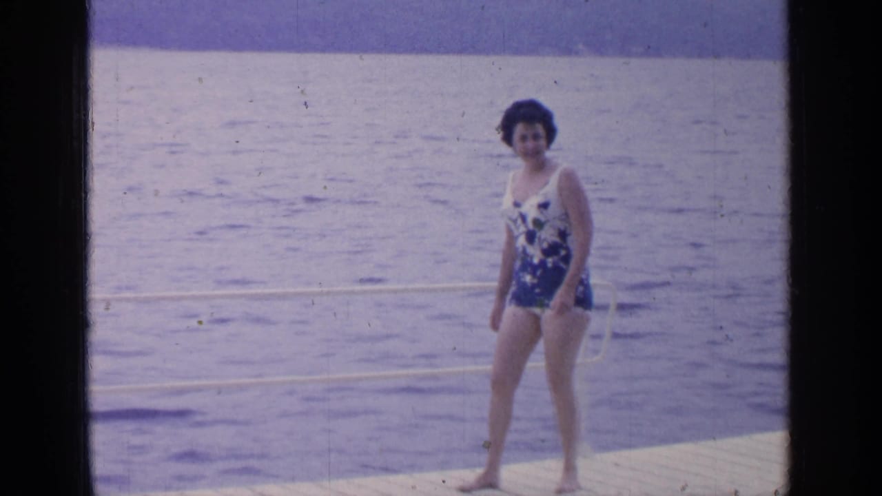 Woman in a floral swimsuit stands barefoot on a wooden dock beside lake water.