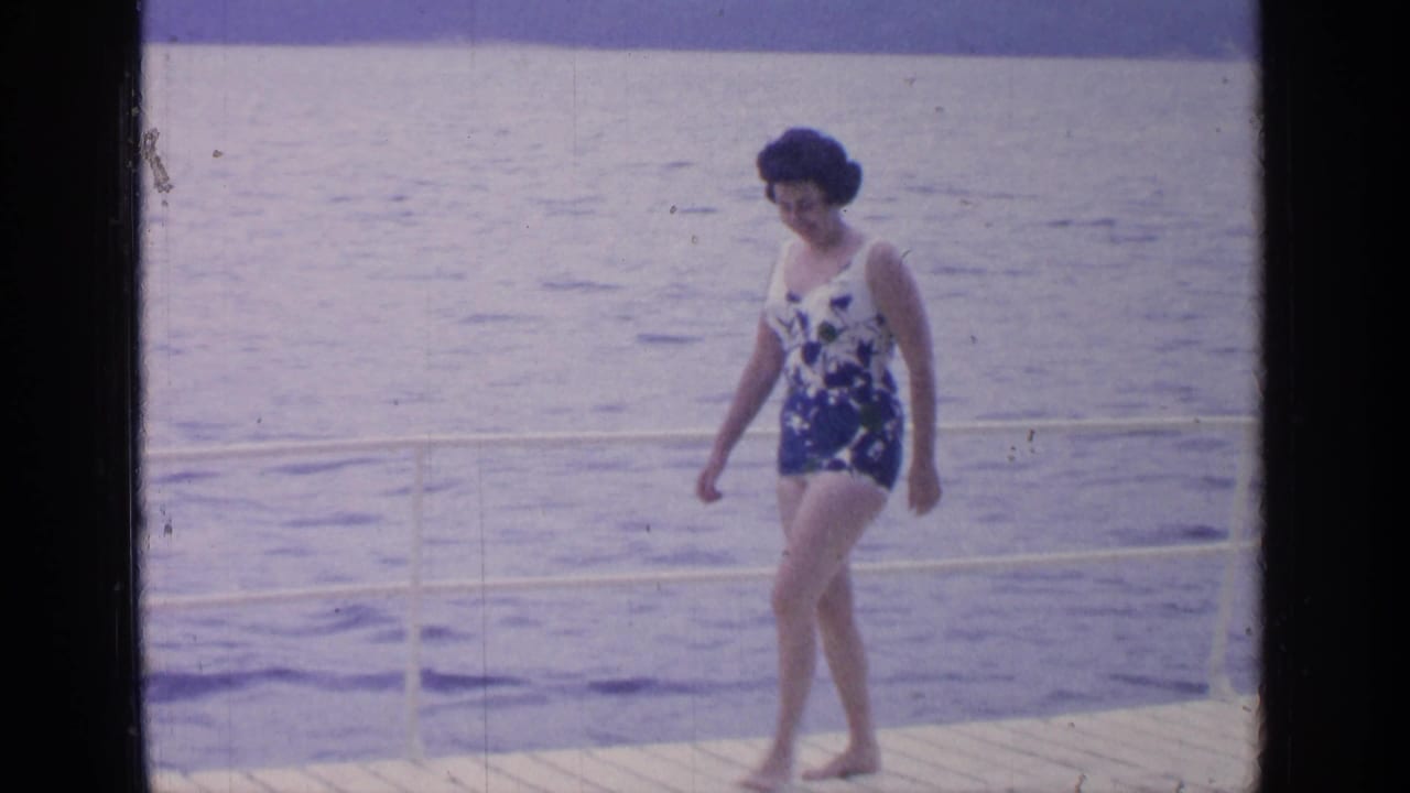 The woman walks barefoot along the Greenwood Lake dock, passing a white railing with the lake filling the background.