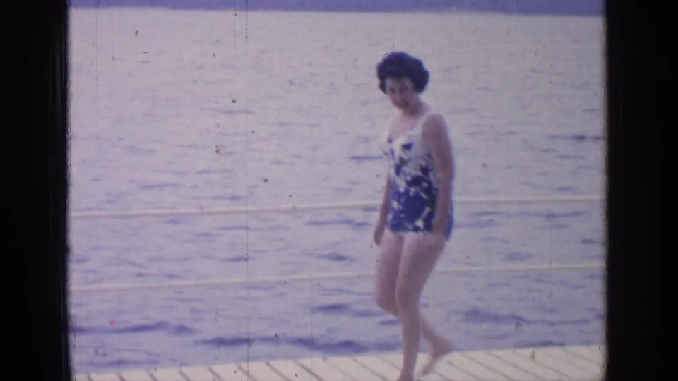 The 1964 home-movie frame shows the woman mid-step on the dock at Greenwood Lake, with soft 8mm color and water in the background.