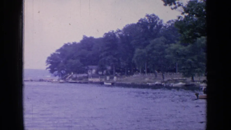 A distant Greenwood Lake shoreline view shows calm water, a wooded bank, small lakeside structures, and the edge of the dock area.