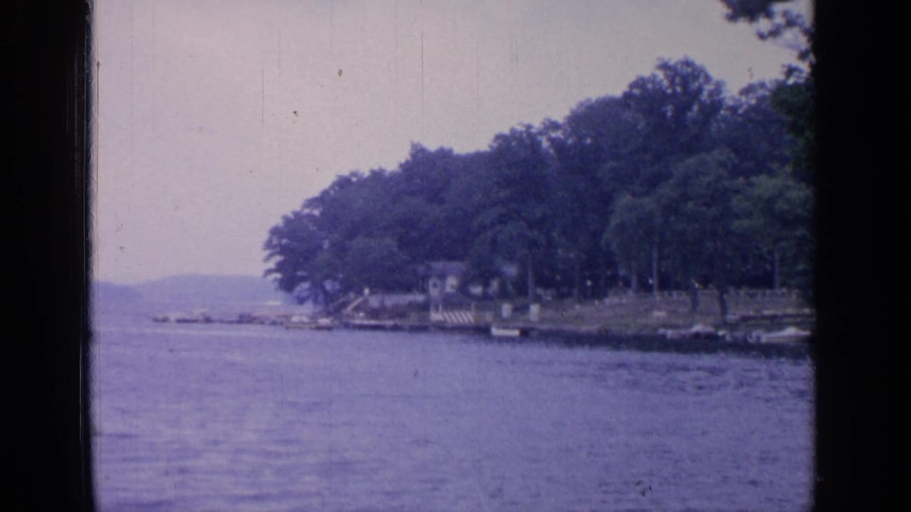 1964 8mm view of Greenwood Lake with calm water, a tree-lined shore, distant hills, and small shoreline structures.