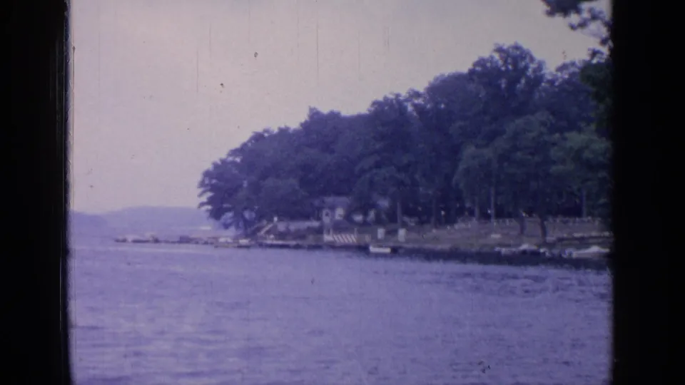 1964 8mm view of Greenwood Lake with calm water, a tree-lined shore, distant hills, and small shoreline structures.