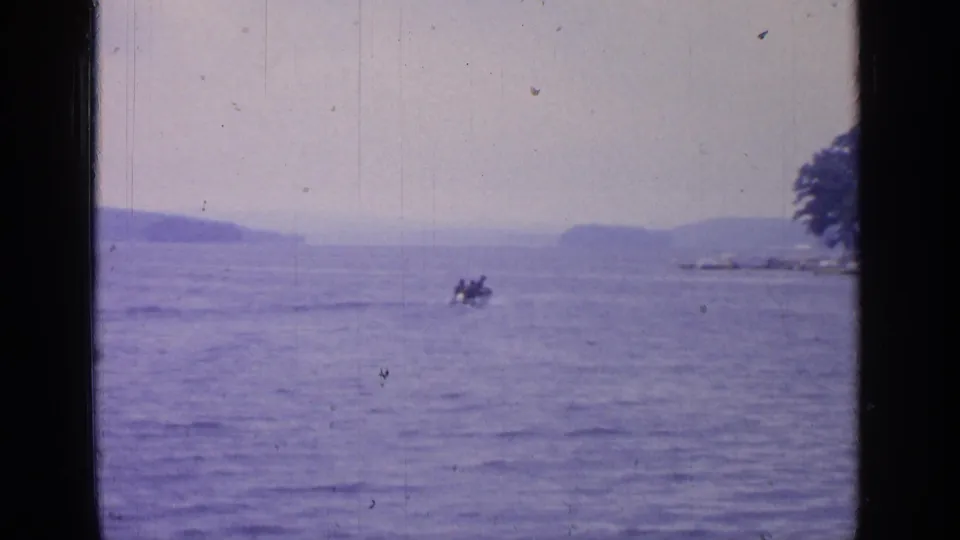 A small motorboat carrying distant figures moves across Greenwood Lake, with wooded shore and low hills in the background.