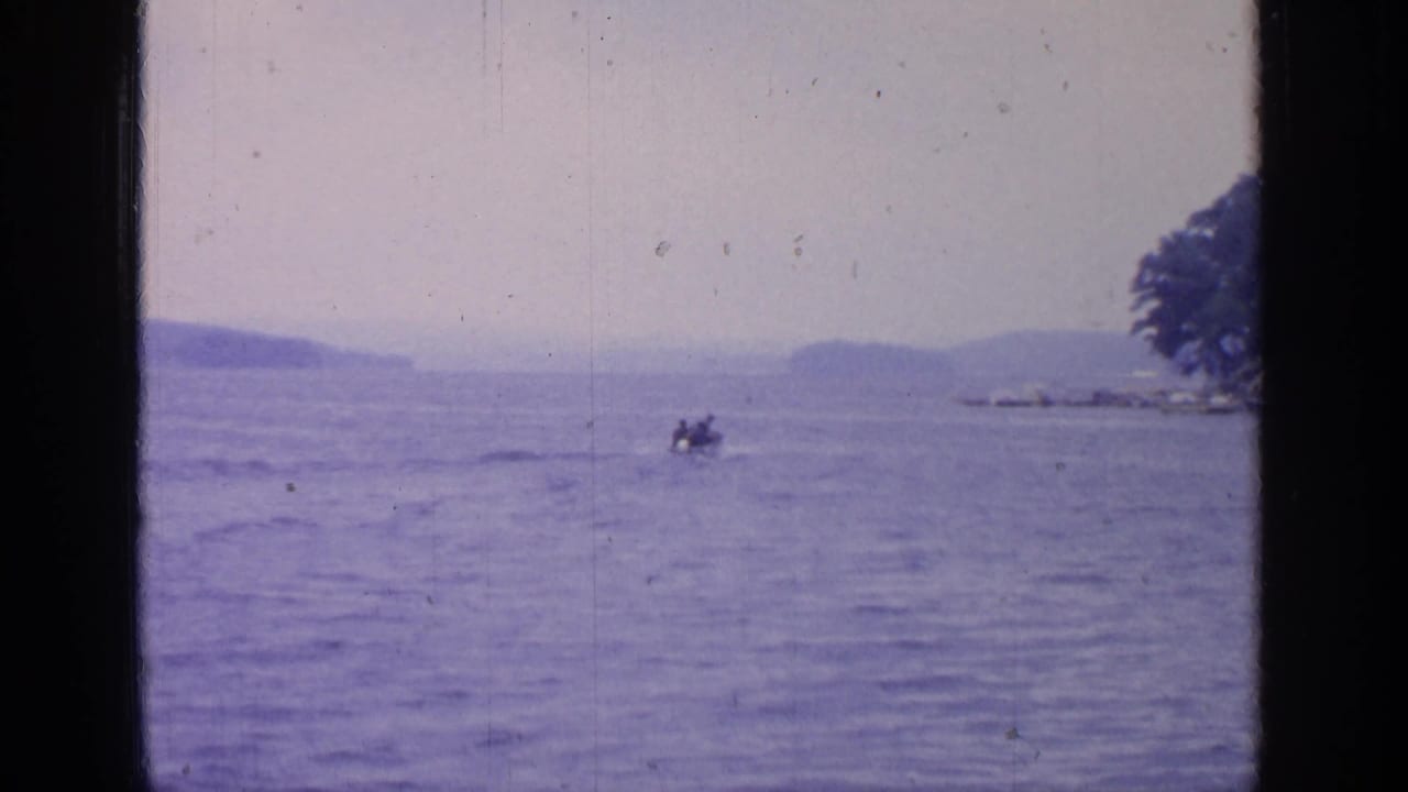 1964 Greenwood Lake view through a dark window frame, showing calm water, distant hills, and a small boat with occupants.