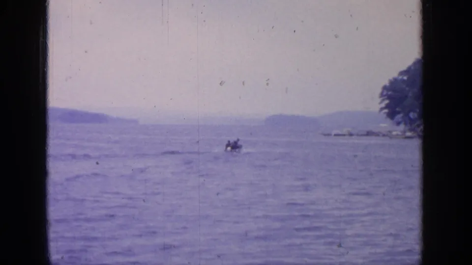 1964 Greenwood Lake view through a dark window frame, showing calm water, distant hills, and a small boat with occupants.