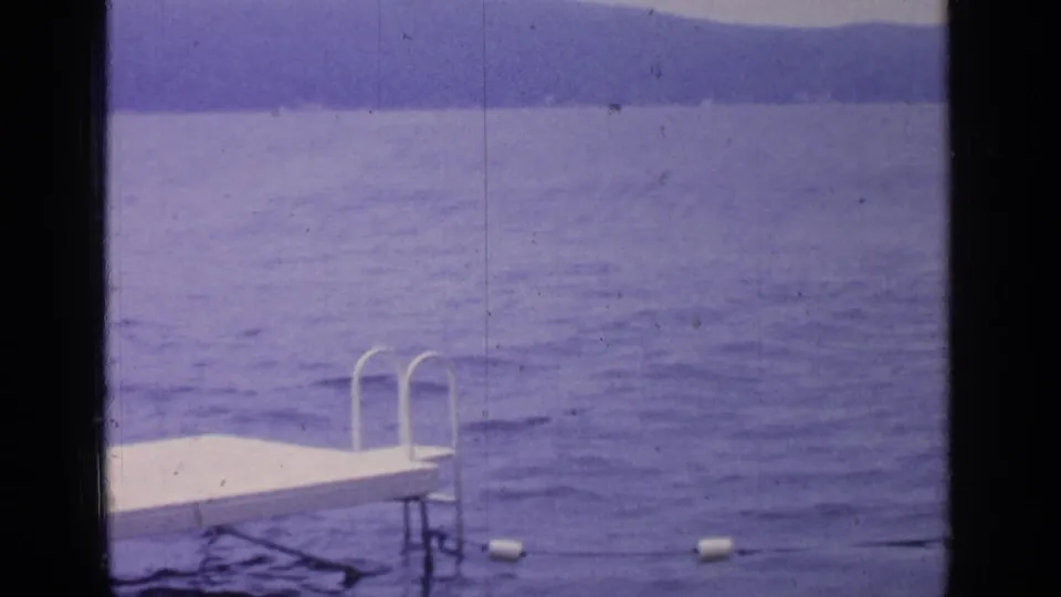 A white lakeside dock with metal ladder rails enters the foreground against Greenwood Lake and hazy mountain ridges.