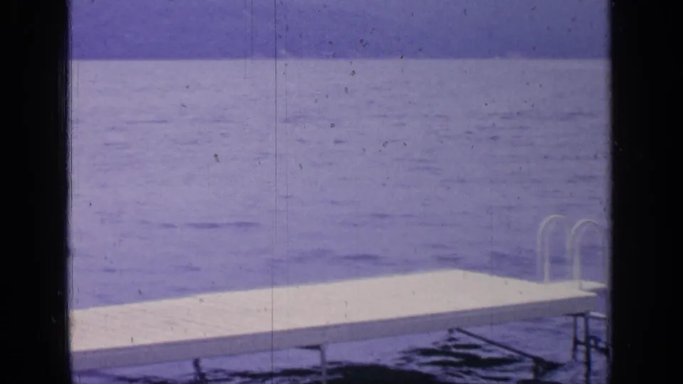 1964 Greenwood Lake home-movie frame showing a white dock, small ladder, calm blue water, and distant shoreline through dark border edges.