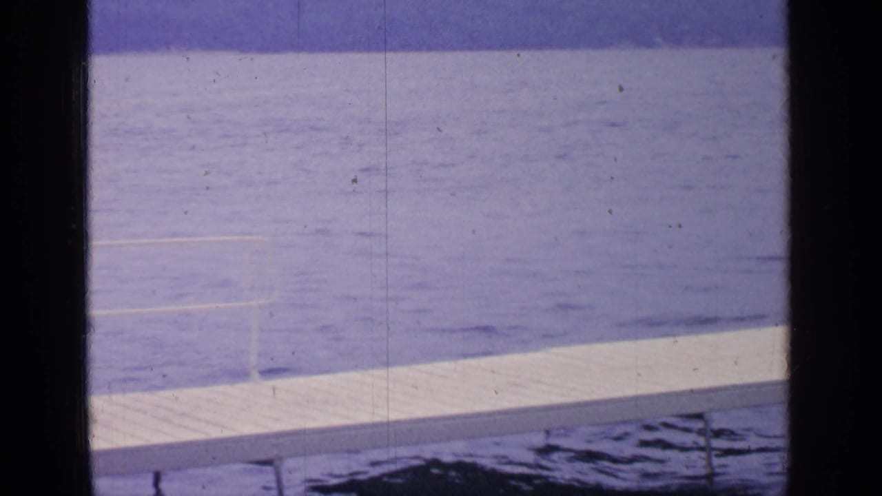 1964 Greenwood Lake frame with a white dock running across blue water, faint ripples, distant shoreline, and dark vertical film edges.