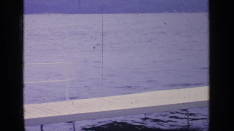 1964 Greenwood Lake frame with a white dock running across blue water, faint ripples, distant shoreline, and dark vertical film edges.