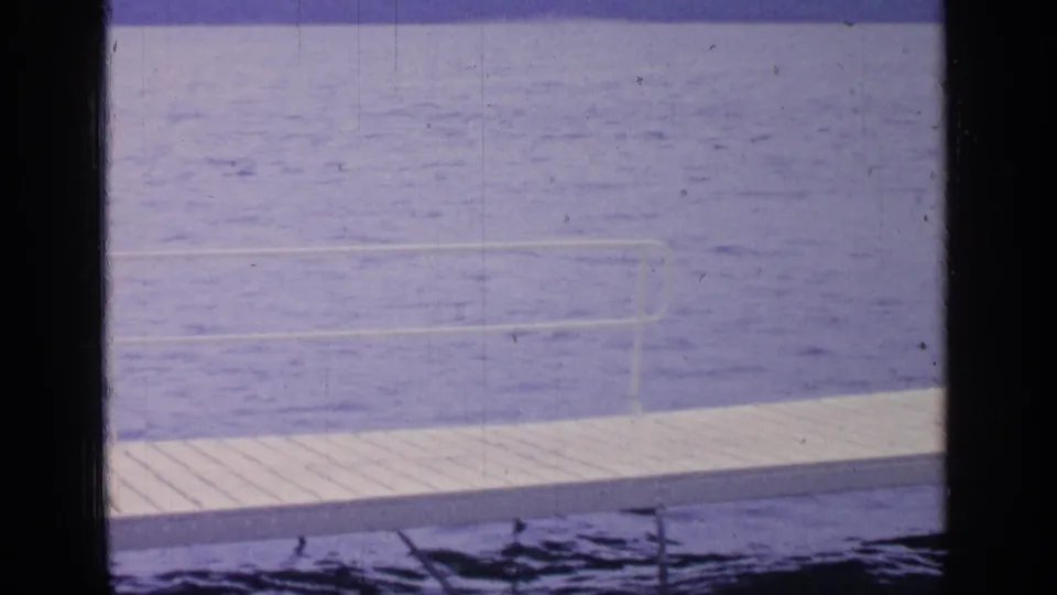 1964 Greenwood Lake home-movie frame looking past dark window-like edges toward blue water, a white dock, and faint railing or ladder forms.