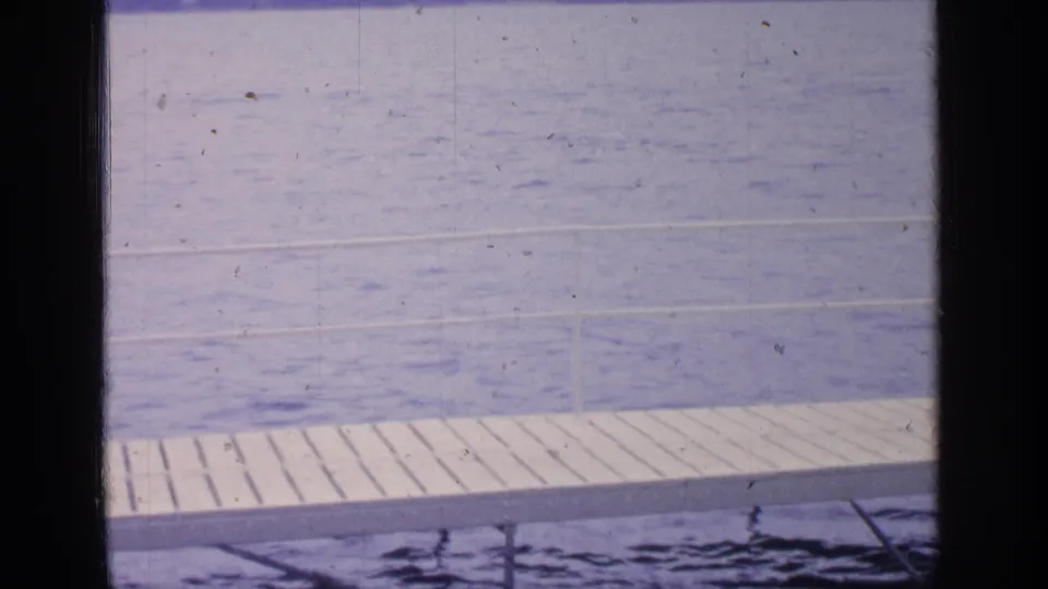 1964 Greenwood Lake frame showing a white plank dock and supports at the lower edge, with calm blue water and faint shoreline beyond.
