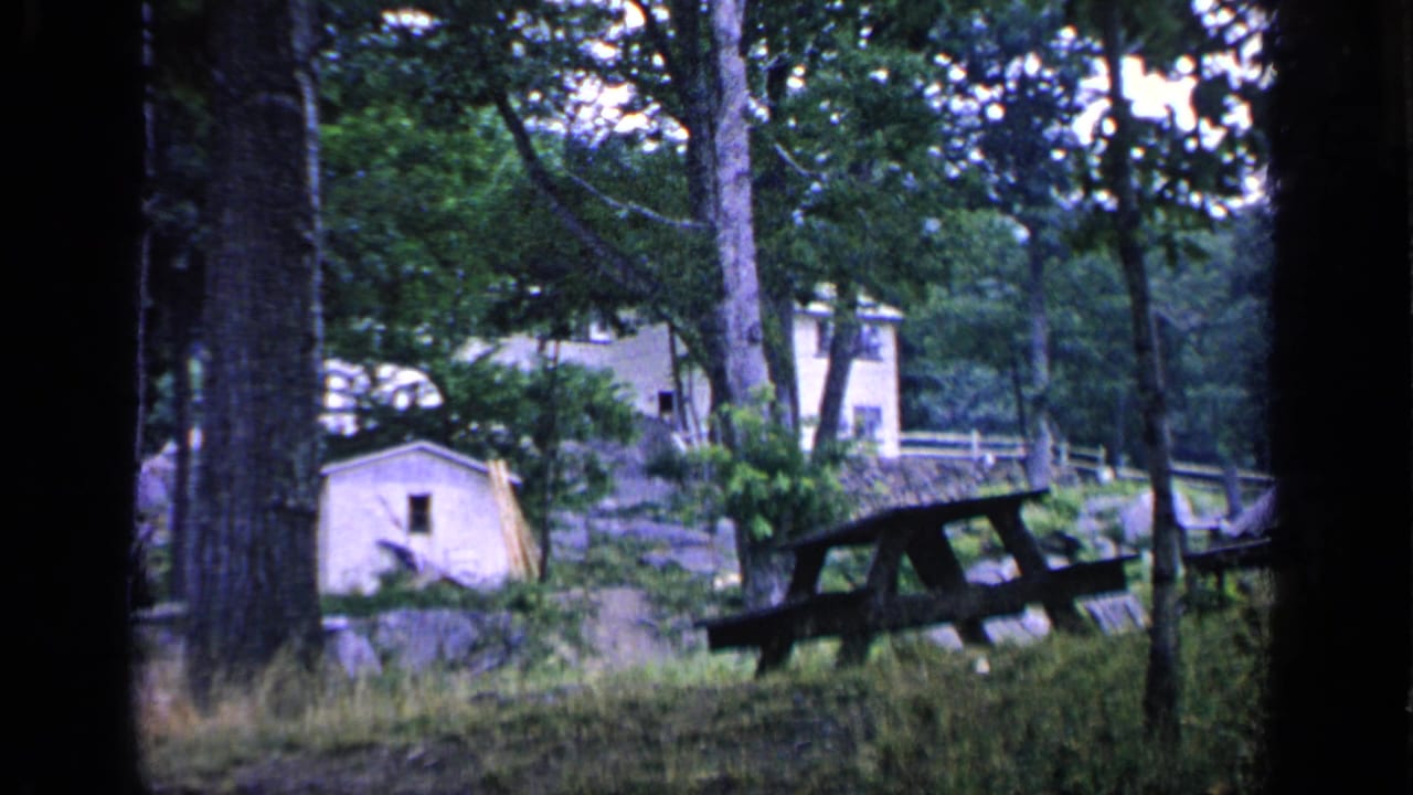 1964 Greenwood Lake home-movie frame showing a shaded wooded clearing with a picnic table, small cabin, trees, grass, and a distant house.