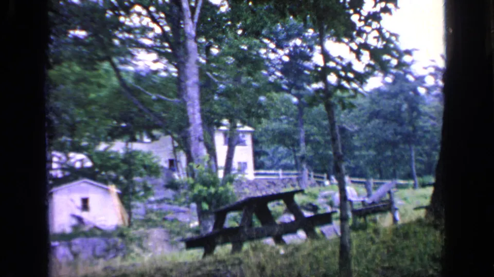 1964 Greenwood Lake home-movie frame showing a grassy wooded picnic area with a pale cabin, larger house, fence, and table.