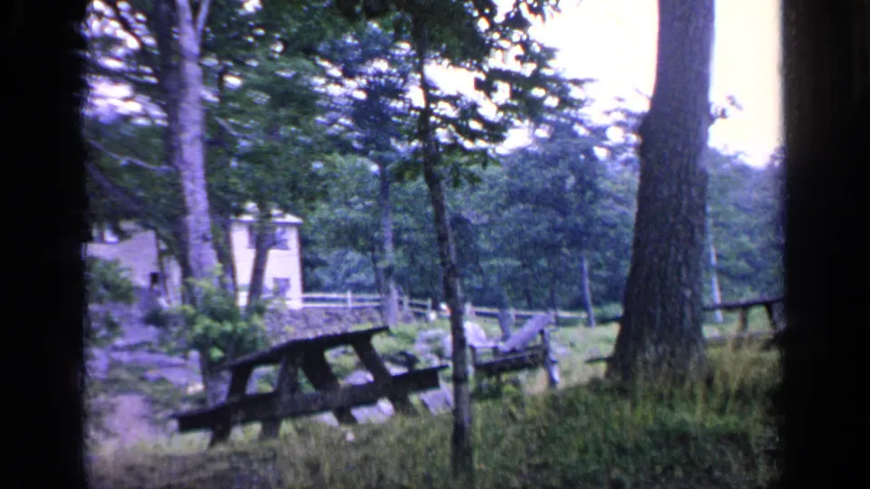 1964 Greenwood Lake frame showing a weathered picnic table under trees, with tall grass, a small cabin, fence, and house beyond.
