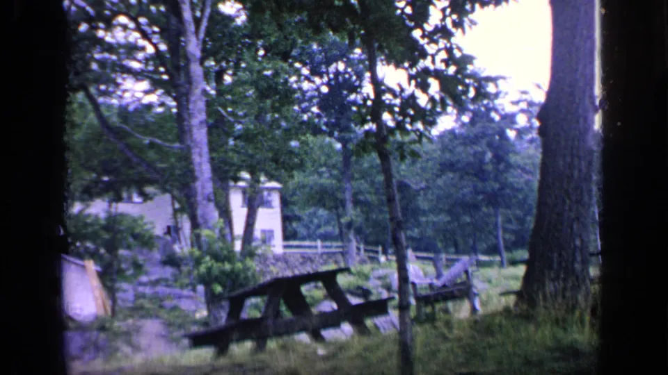 1964 Greenwood Lake frame looking through trees toward weathered picnic tables and a small pale house or outbuilding in the woods.