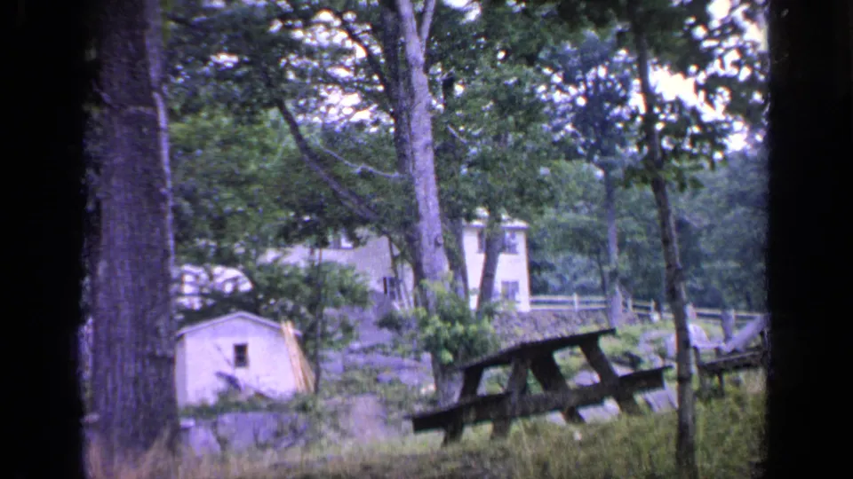 1964 Greenwood Lake 8mm frame with a small shed, weathered picnic table, tall trees, and dense summer foliage.