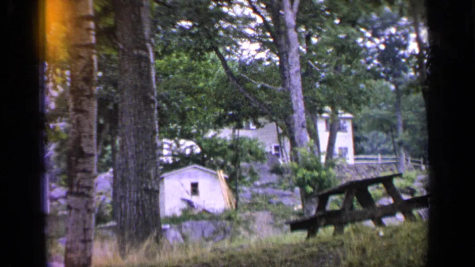 1964 Greenwood Lake frame showing a wooded picnic area, small shed, tall trees, and a house partially hidden by leaves.
