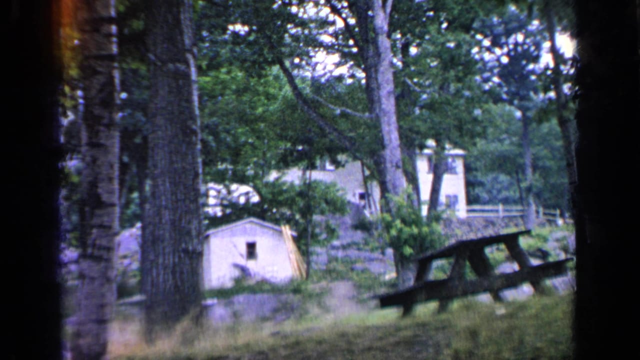 Greenwood Lake home-movie frame showing trees, a small shed, a picnic table, and cabin grounds in a wooded summer setting.