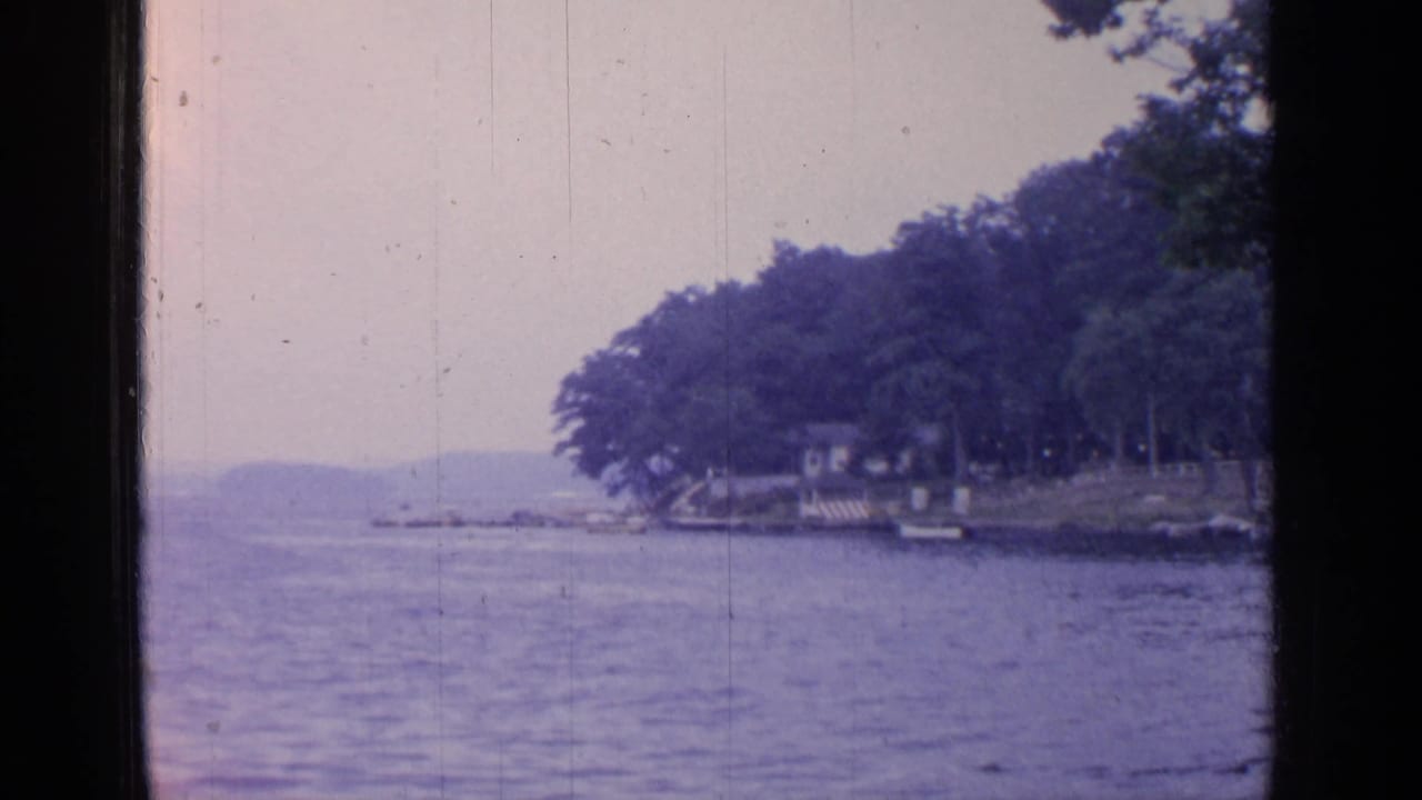 Greenwood Lake view with calm water, wooded shoreline, small boats, and distant hills in soft 8mm color.