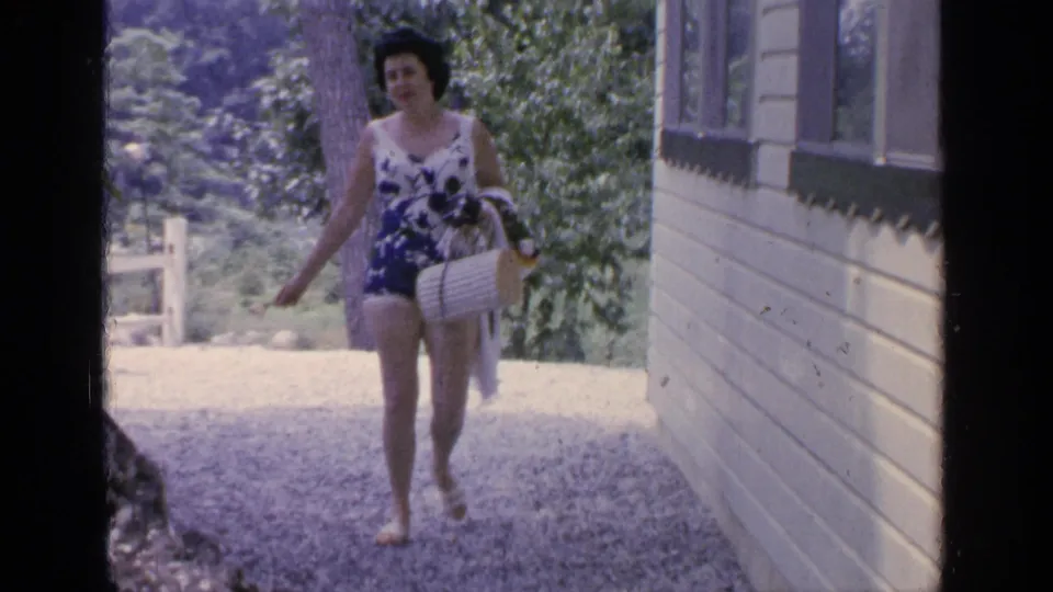 Middle-aged woman at Greenwood Lake in 1964 walks barefoot on gravel beside a cabin, carrying a wicker basket in summer attire.