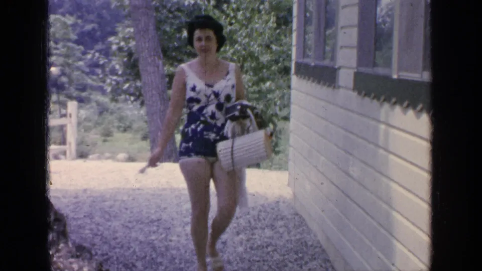 Woman in a blue and white floral swimsuit approaches the camera along a lakeside cabin path in 1964 Greenwood Lake footage.