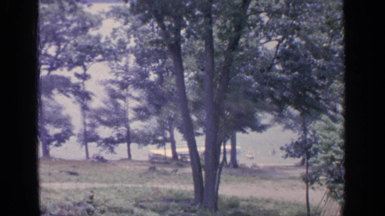 1964 Greenwood Lake home-movie frame showing trees, grass, bright water, and a distant shoreline view through dark vertical borders.