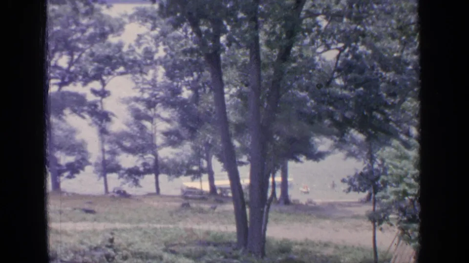 1964 Greenwood Lake home-movie frame showing trees, grass, bright water, and a distant shoreline view through dark vertical borders.