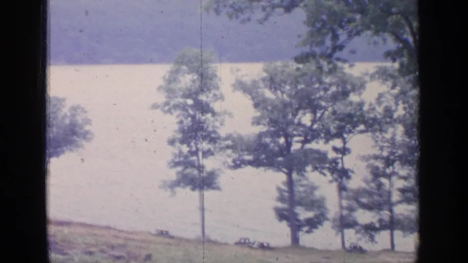 1964 lakeside park view at Greenwood Lake, with trees, open grass, bright water, and small distant cars along the shore.