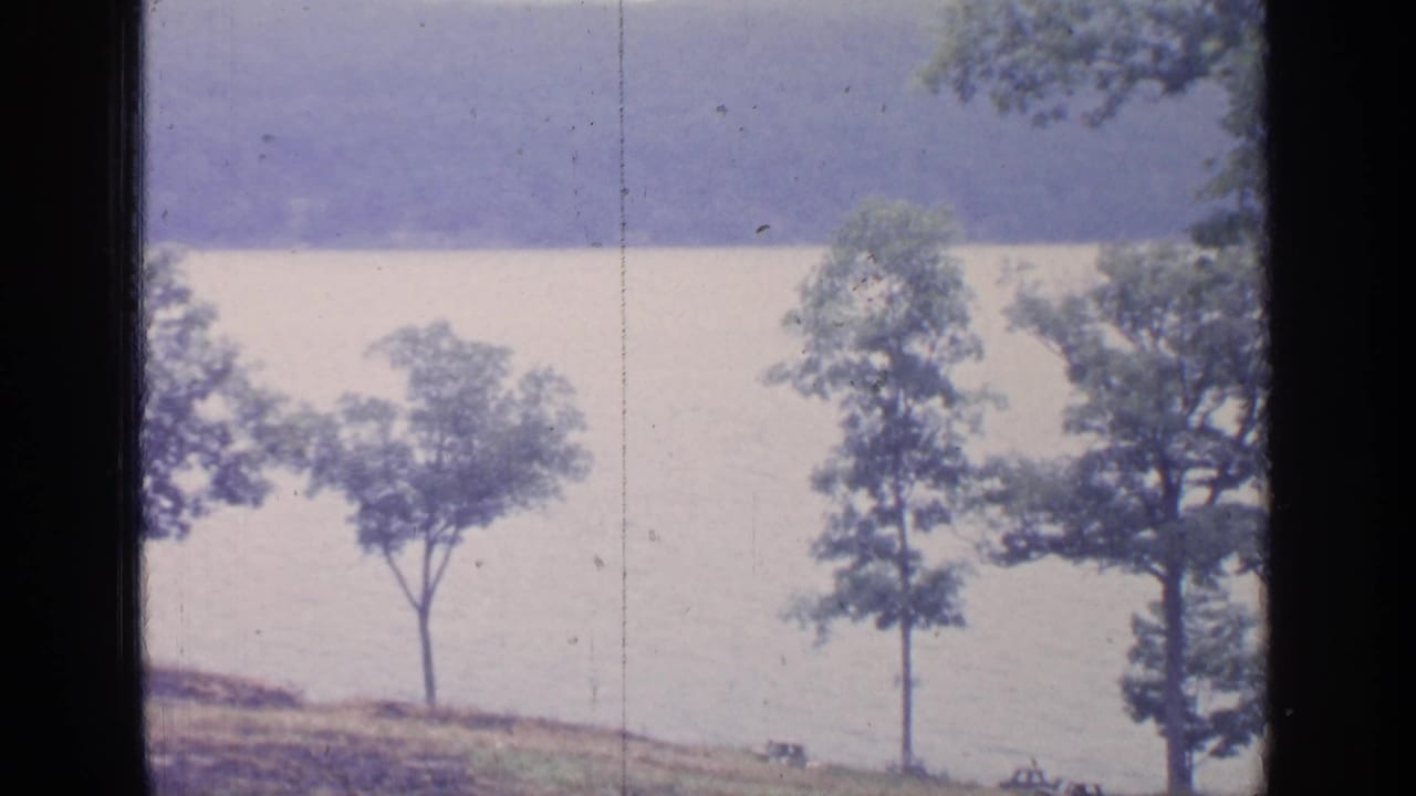 1964 Greenwood Lake frame with shoreline trees, calm water, distant hills, and small dark shapes near the lower shore.