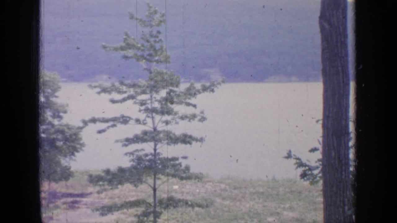 1964 8mm view of Greenwood Lake, New York, seen through dark window framing with young trees in the foreground and wooded hills beyond.