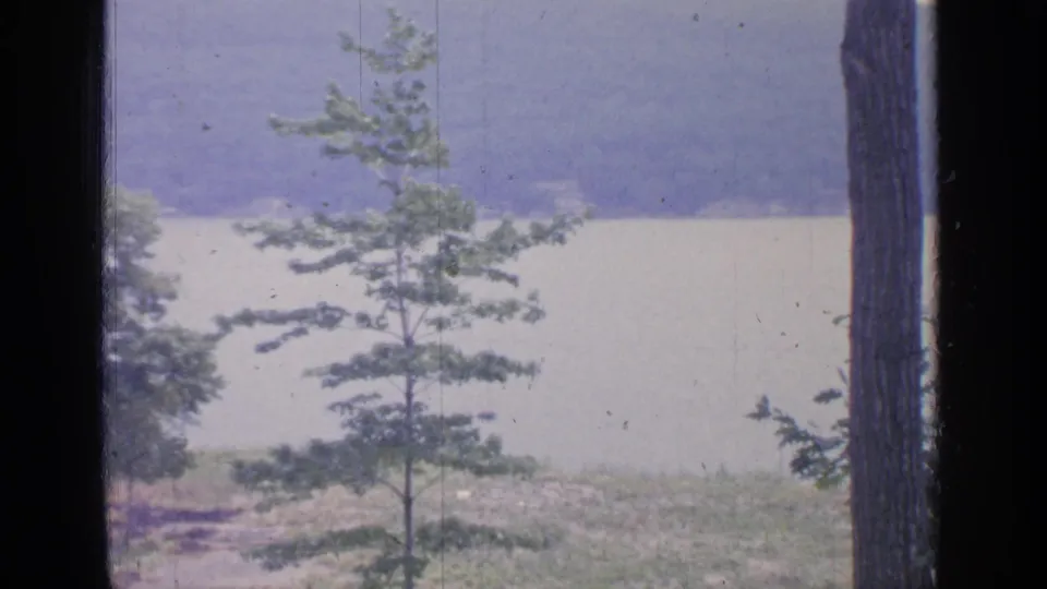 1964 8mm view of Greenwood Lake, New York, seen through dark window framing with young trees in the foreground and wooded hills beyond.