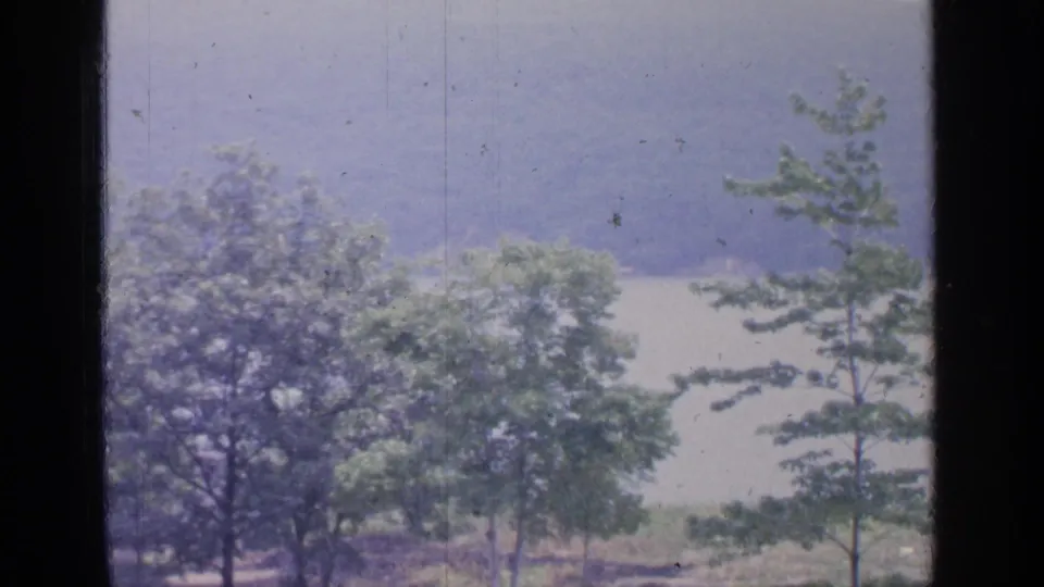 Final 8mm frame of the Greenwood Lake view, with trees and shrubs near the shore, calm water behind them, and muted wooded hills in the distance.