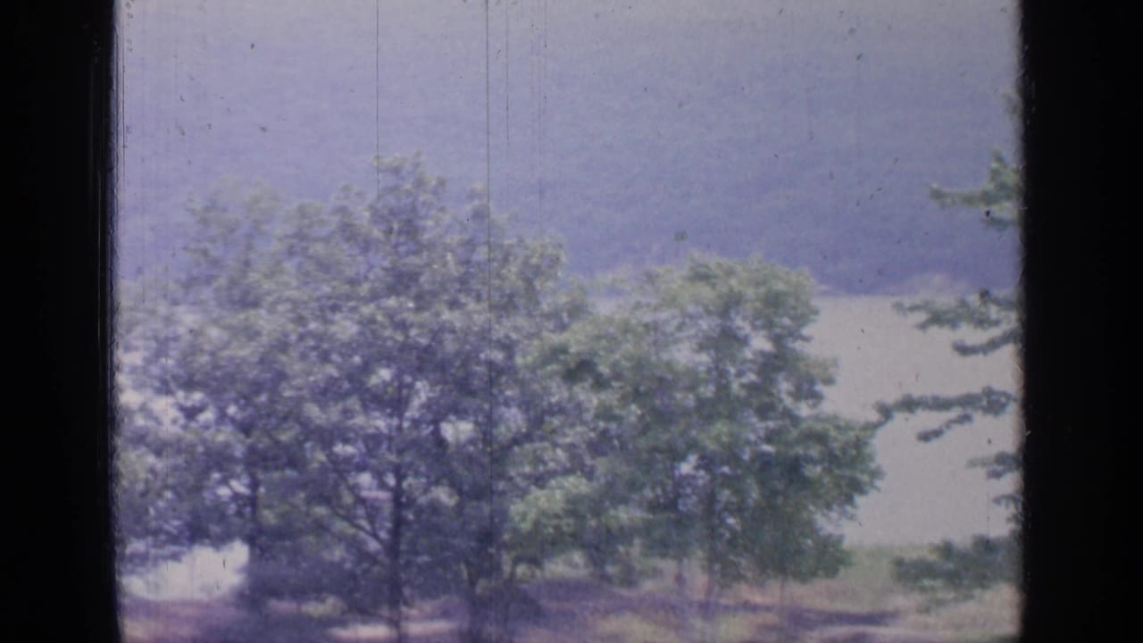 1964 Greenwood Lake home-movie view through a window toward trees, pale water, and hazy hills beyond.