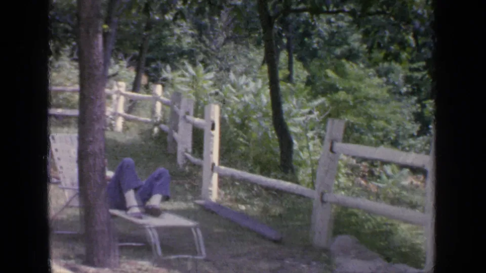 In a wooded Greenwood Lake yard, a man reclines on a lawn chair with his feet up near a split-rail fence and greenery.
