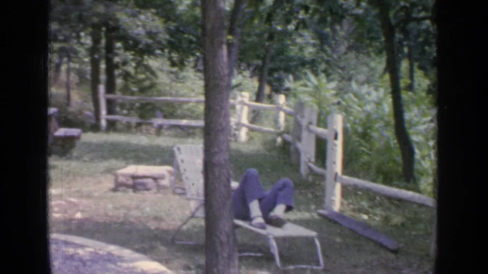 The man remains reclined in a lawn chair, with a split-rail fence, picnic table, trees, and dense greenery around him.